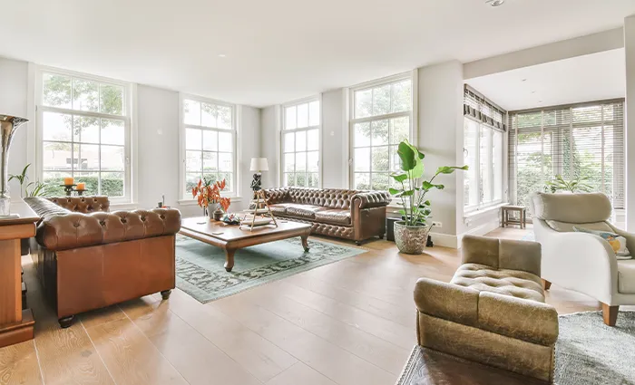 Salon lumineux avec canapés en cuir marron, table basse en bois, plantes vertes et grandes fenêtres, illustrant un intérieur spacieux et élégant.